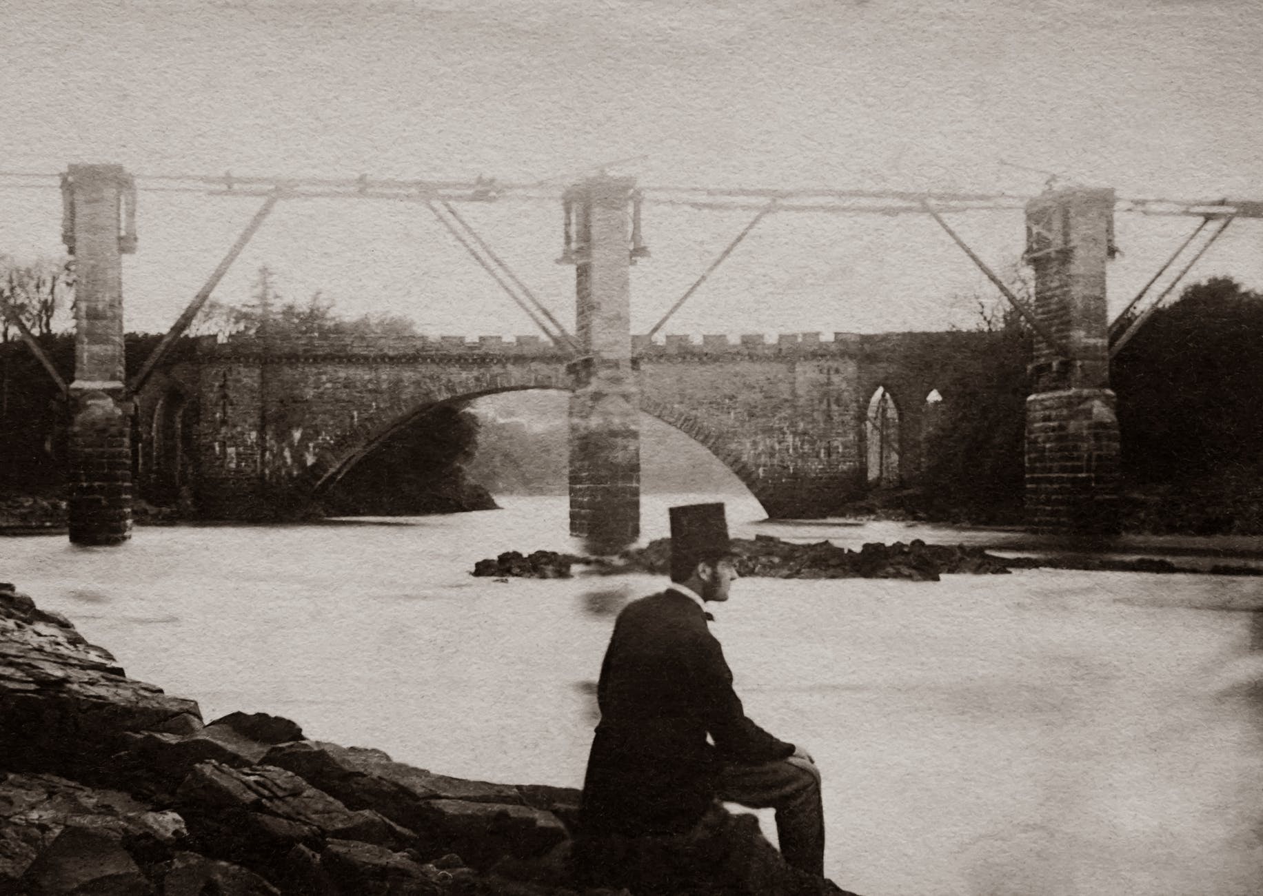 grayscale photo man wearing a top hat and suit sitting by the river near a bridge