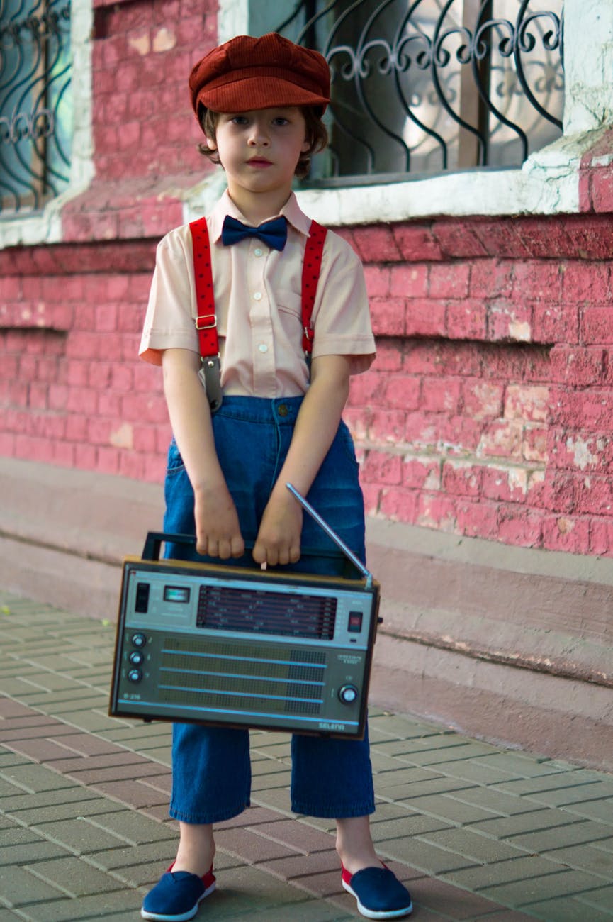 photo of boy holding radio and standing beside of red house
