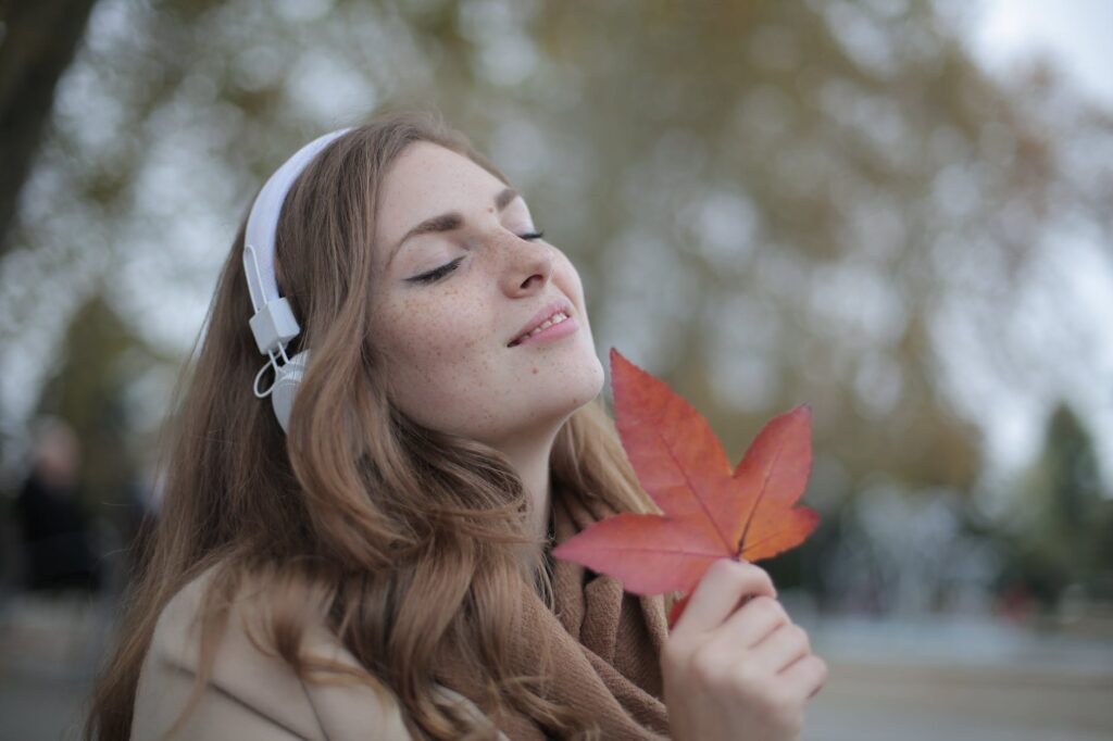 young satisfied woman in headphones with fresh red leaf listening to music with pleasure while lounging in autumn park