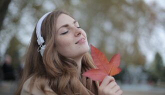 young satisfied woman in headphones with fresh red leaf listening to music with pleasure while lounging in autumn park